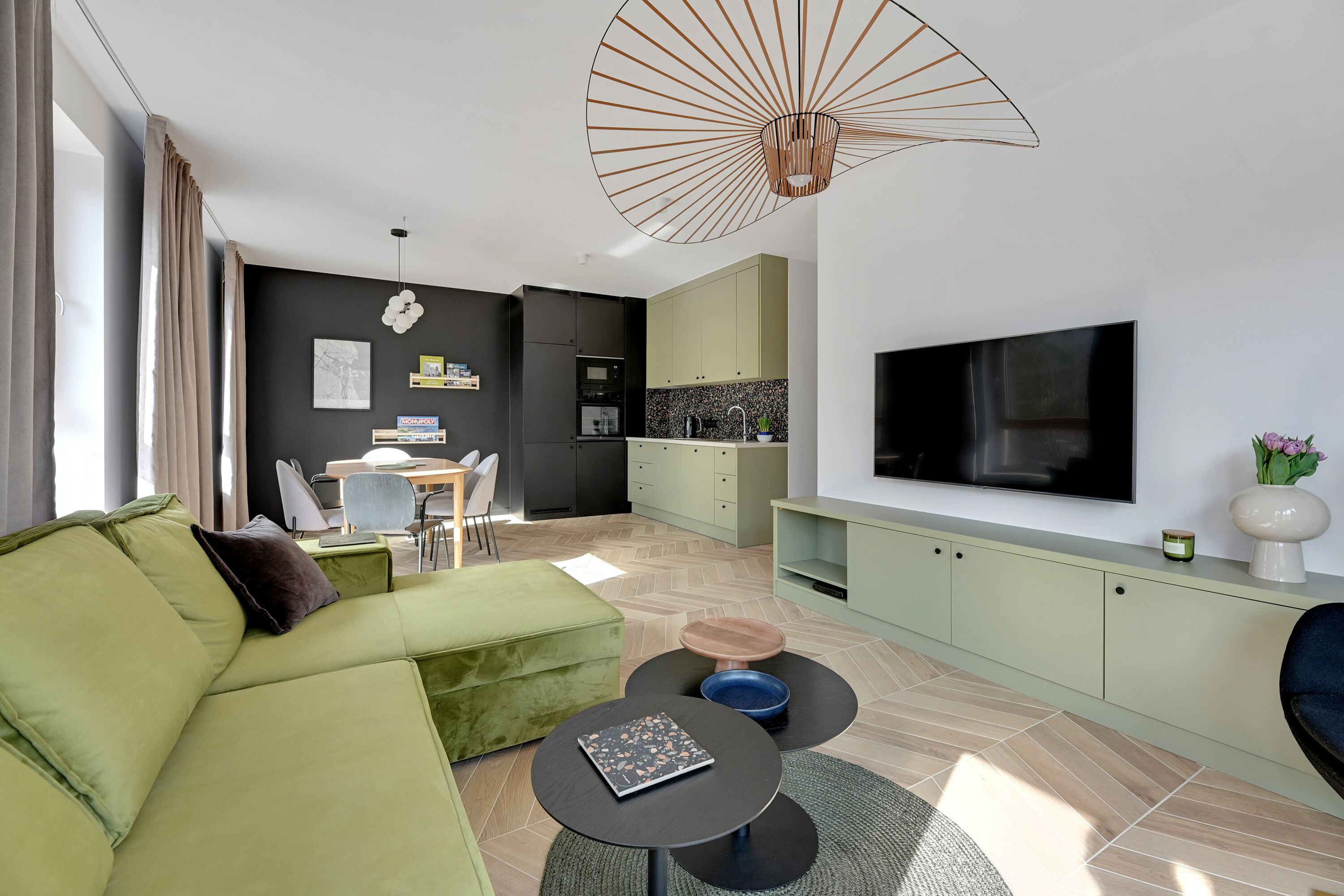 A modern living room with a green sofa, a dining area with a table and chairs, and a kitchen with a TV above the counter.