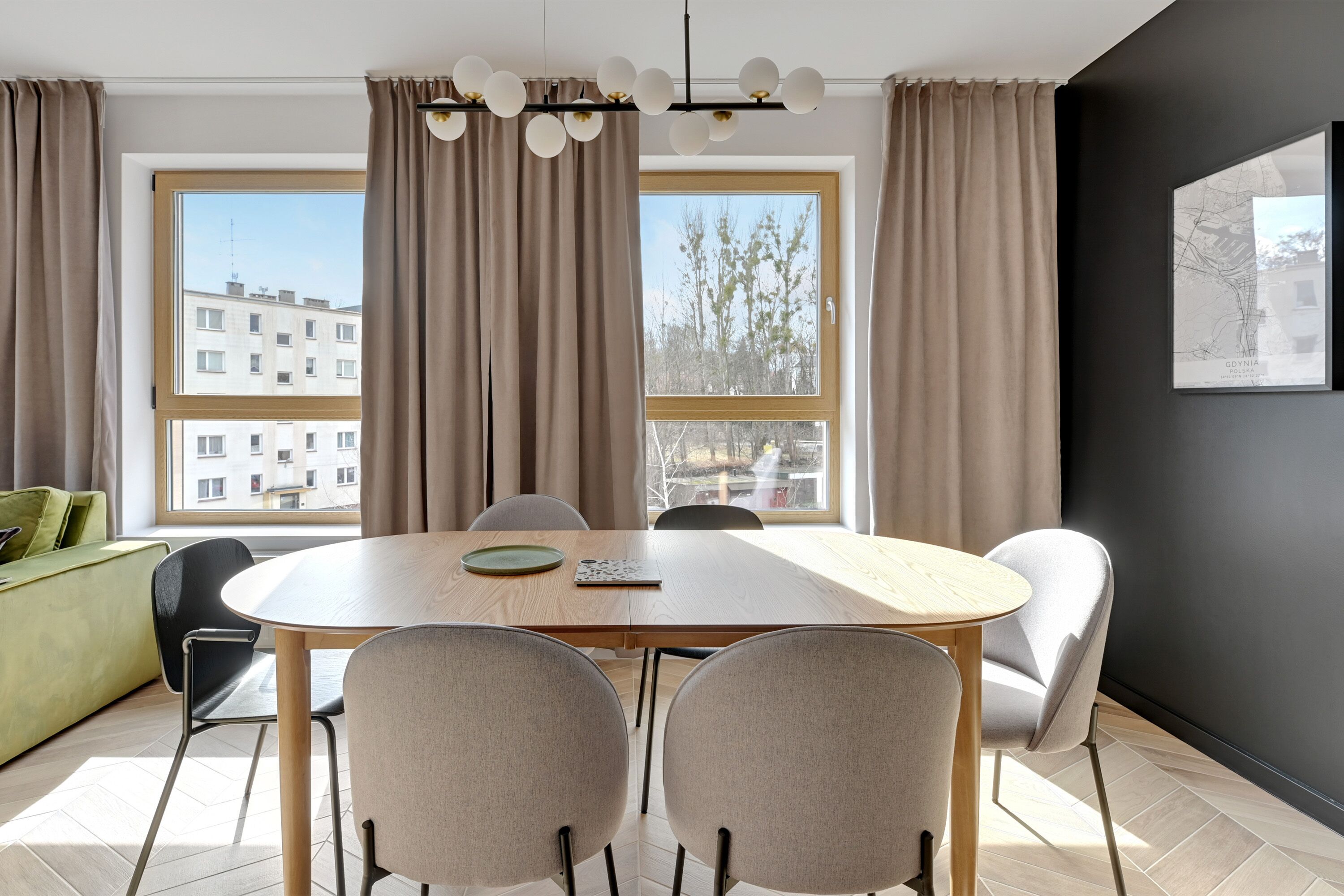 A modern dining area with a wooden table and four chairs, large windows with curtains, a green sofa, and a black wall with a framed picture.