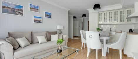 A modern living room with a sofa, coffee table, and dining area. There are framed pictures on the wall and a kitchen with white cabinets.