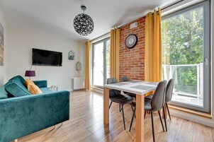 A modern living room with a teal sofa, a dining table with chairs, a TV mounted on the wall, and a large window with curtains.