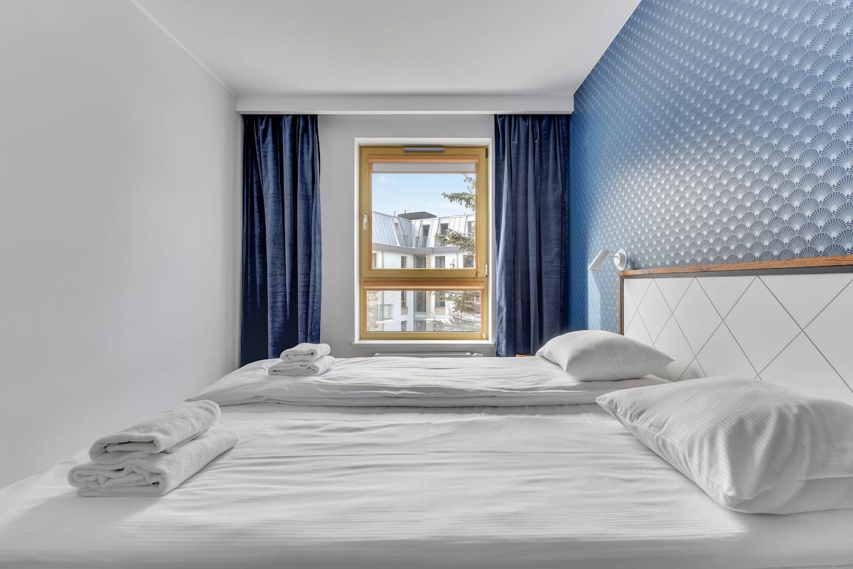 Een net opgemaakt bed met witte lakens en kussens, een raam met blauwe gordijnen, en een blauwe, gestructureerde muur.