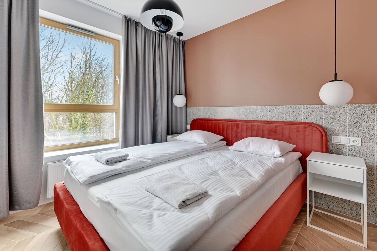 Een net opgemaakt tweepersoonsbed met witte lakens, een rood hoofdbord en een witte nachtkastje.