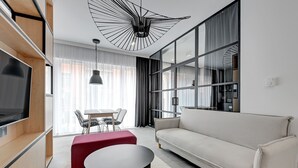 Un salon moderne avec un canapé, un pouf rouge, une table basse ronde et un téléviseur posé sur une étagère en bois.