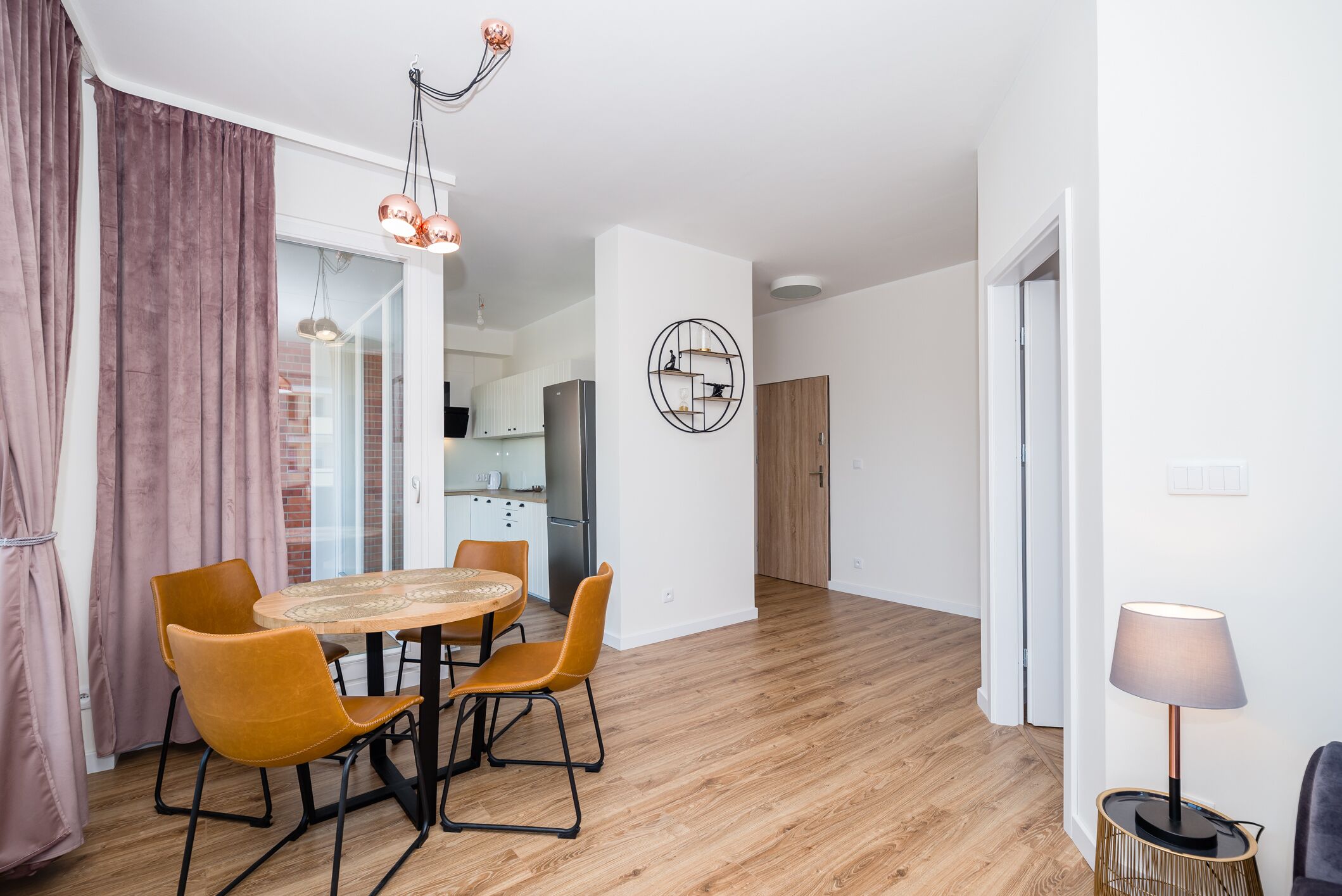 Una sala moderna con una mesa de comedor de madera, dos sillas, una lámpara y una cocina al fondo.