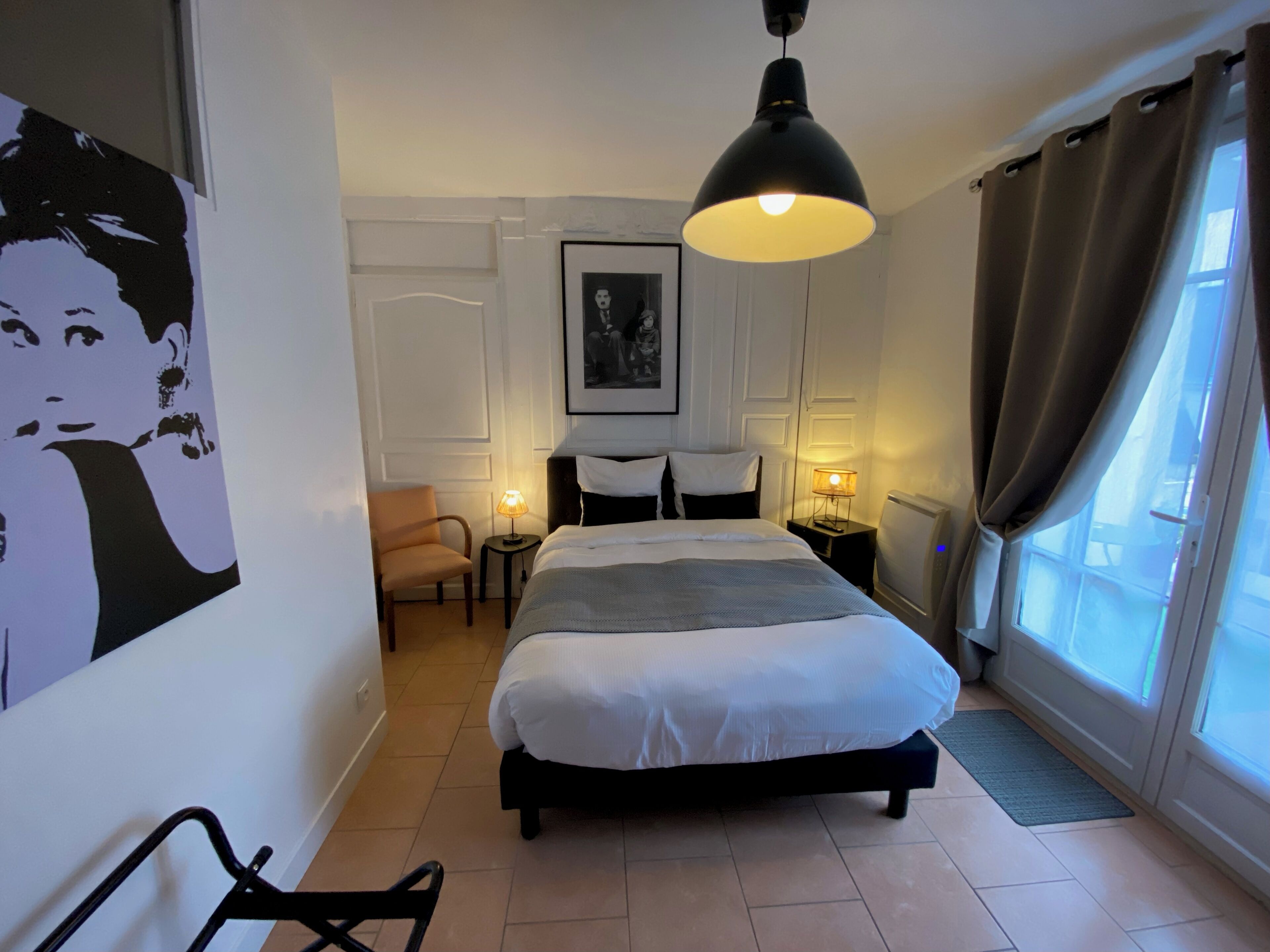 A hotel room with a bed, a chair, a desk with a lamp, a picture on the wall, and a window with curtains.