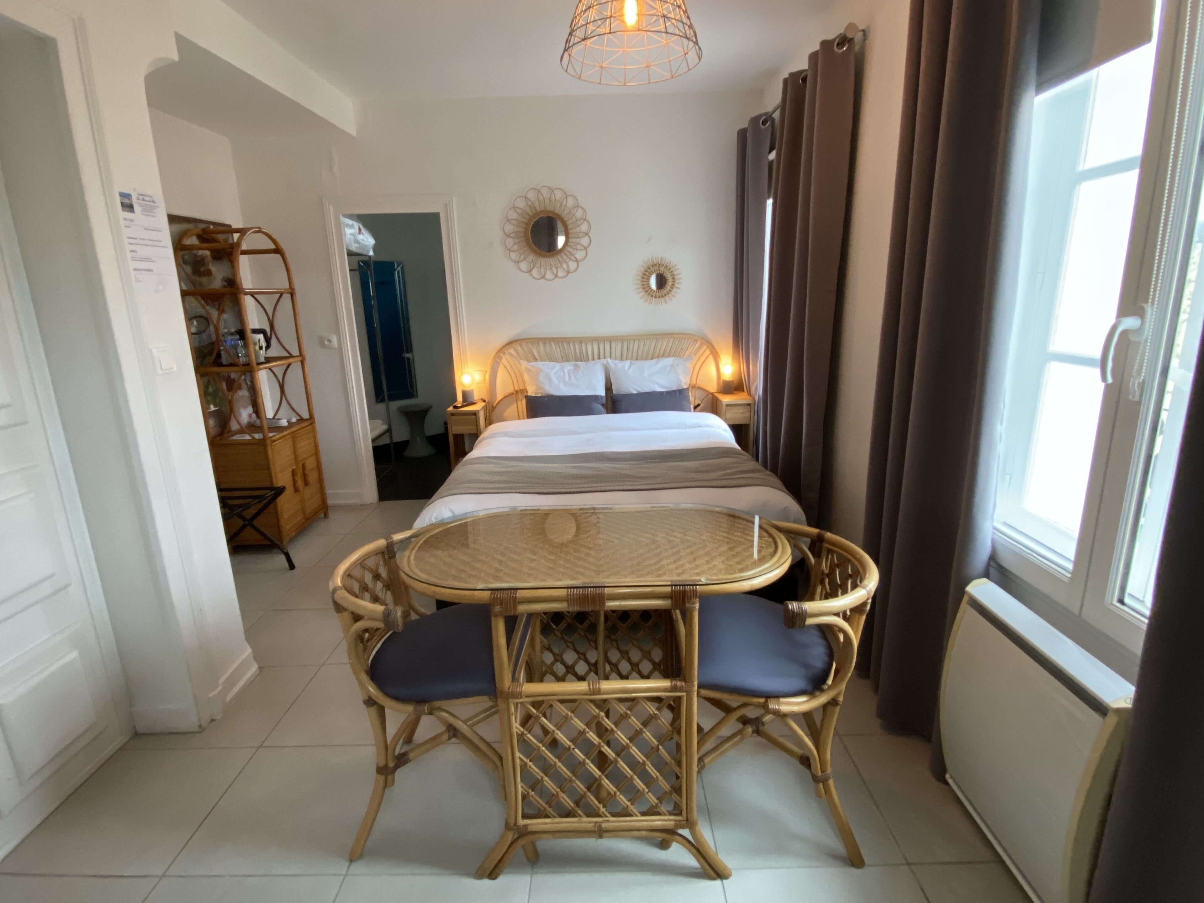 Un dormitorio con cama, mesitas de noche, una mesa redonda con sillas, un espejo y una ventana con cortinas.