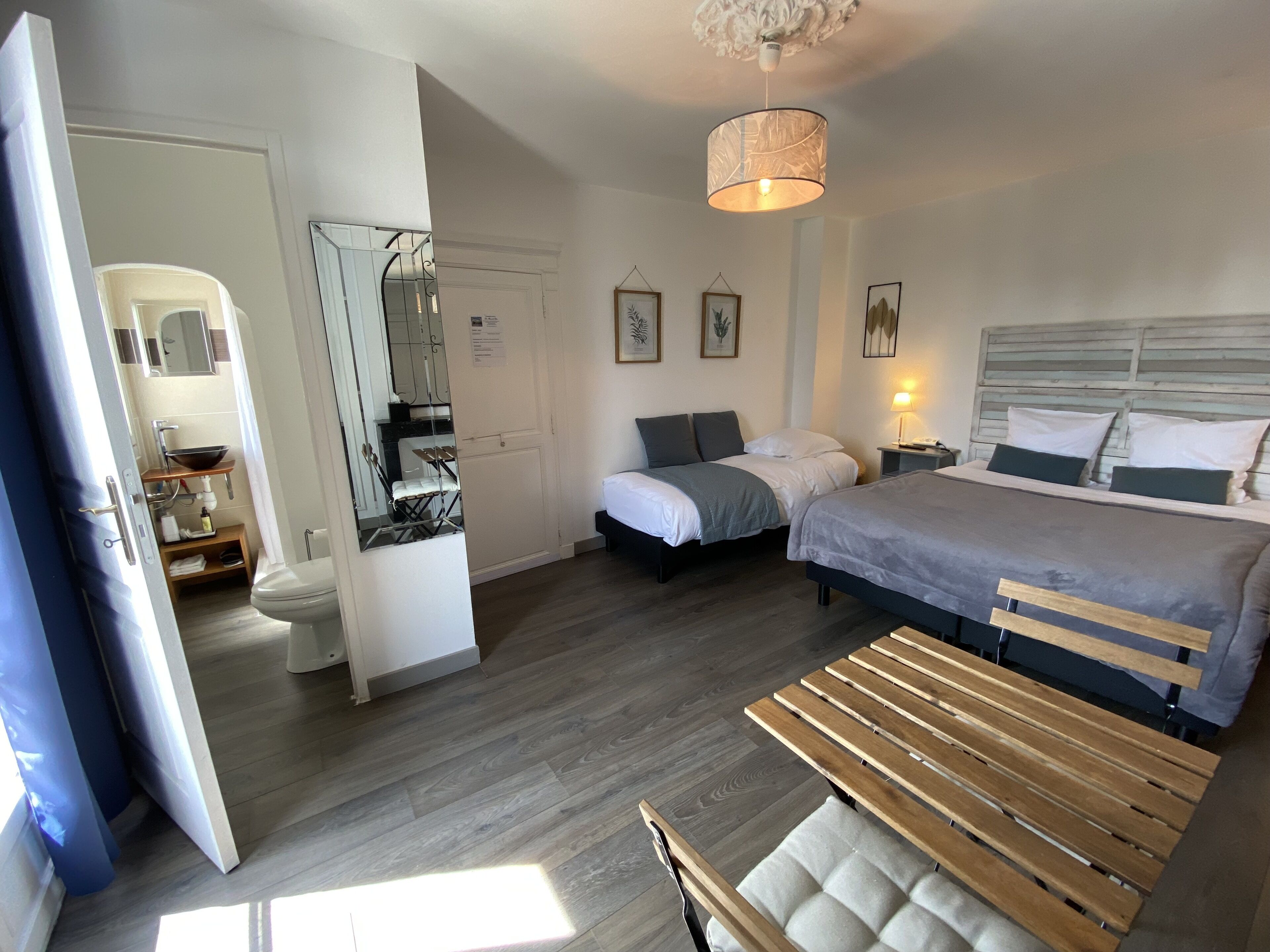 Een slaapkamer met twee bedden, een houten bankje, een plafondlamp en een badkamer met spiegel en wastafel.