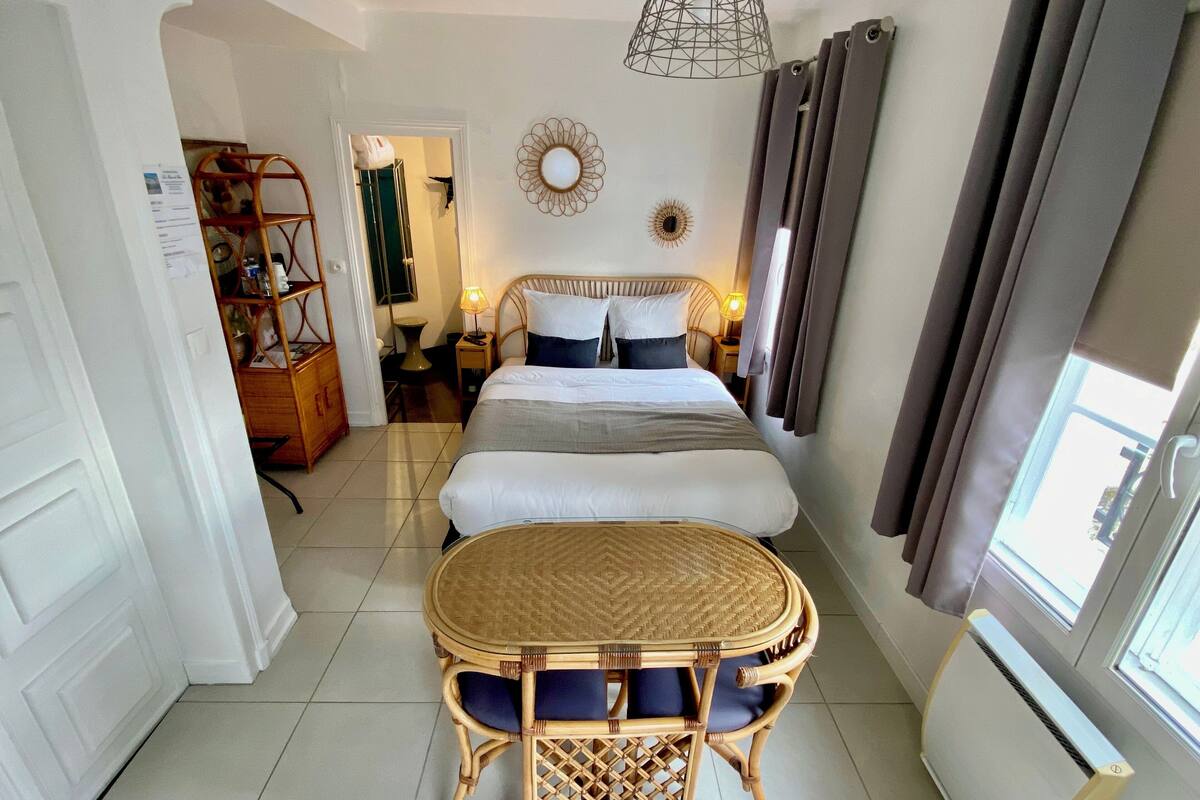 A hotel room with a bed, bedside tables, a wicker table with chairs, a wall-mounted lamp, and a window with curtains.