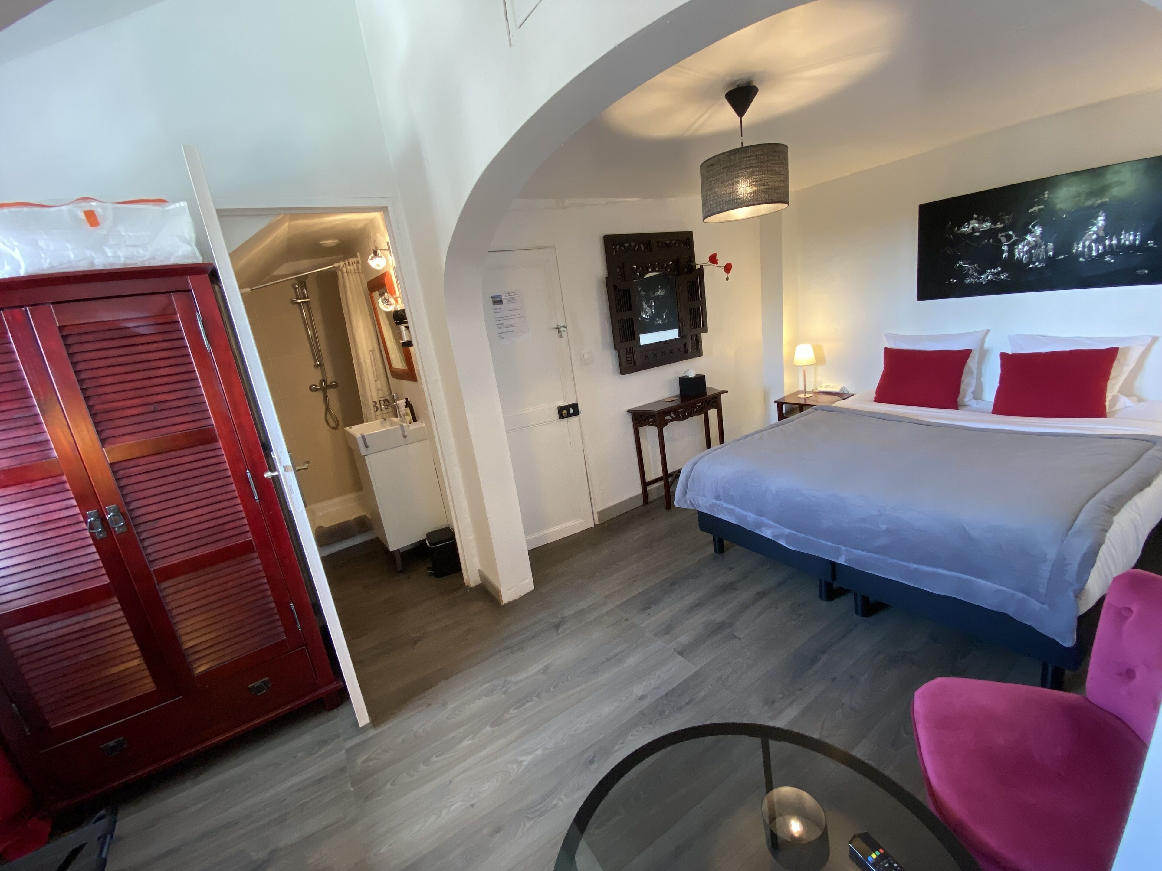 A hotel room with a bed, a desk, a chair, a mirror, a nightstand, and a red door.