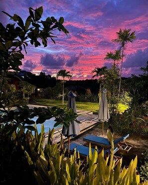 A pool area with lounge chairs and umbrellas, surrounded by tropical plants and palm trees, under a twilight sky. - Druwa Bali (Kutuh)