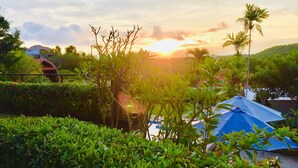 A lush garden with a blue-roofed structure, surrounded by dense greenery and a sunset in the background. - Druwa Bali (Kutuh)