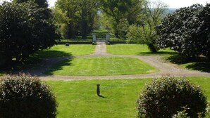 Un jardin bien entretenu, avec un chemin central menant à une construction, le tout entouré d’une végétation luxuriante et d’arbres.