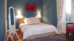 Une chambre à coucher avec un lit, deux lampes de chevet, une œuvre d’art rouge, une fenêtre avec des rideaux et une chaise en bois.