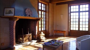 Une pièce avec une cheminée en briques, des poutres en bois, un fauteuil blanc et une table avec des livres.