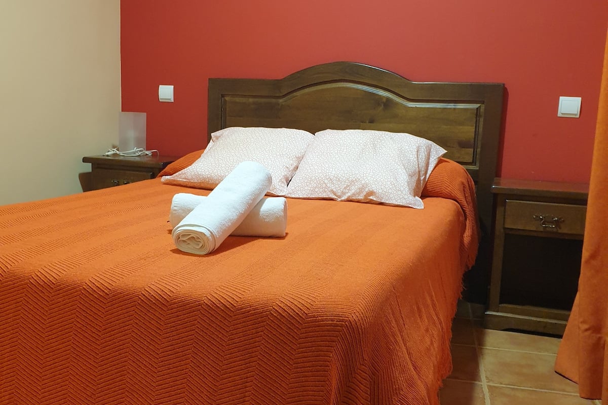 Een net opgemaakt bed met een oranje sprei, twee kussens en een opgerolde witte handdoek.
