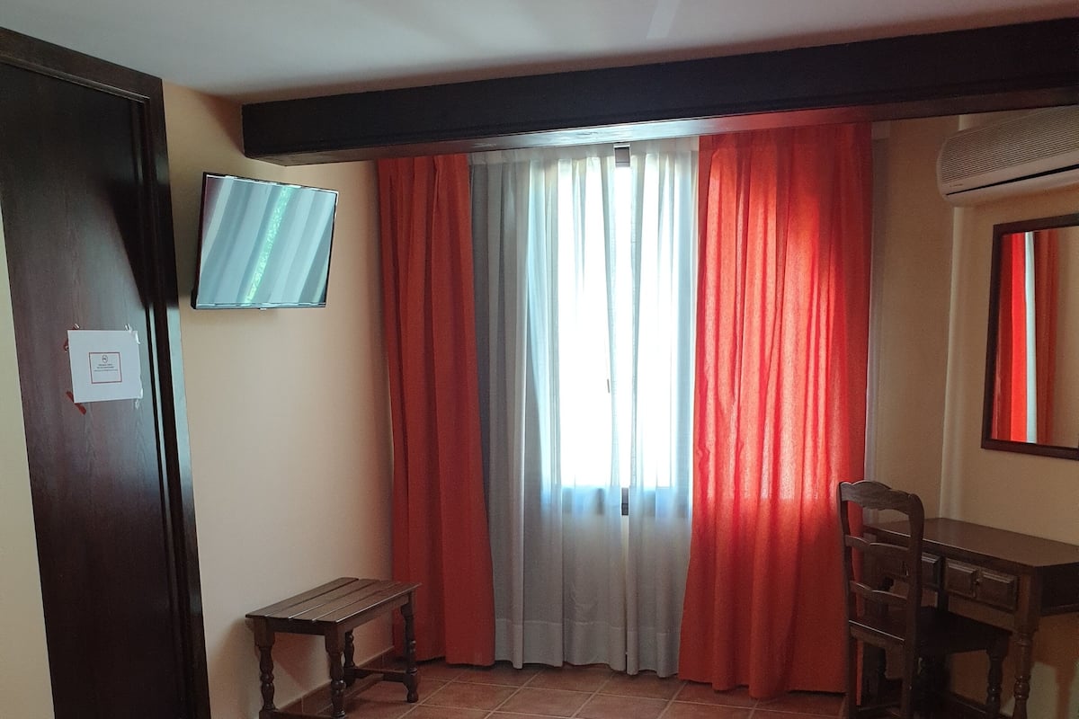 Een slaapkamer met een bed, een bureau, een stoel, een televisie en een raam met gordijnen.