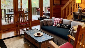 A cozy cabin interior with wooden walls, a sofa with checkered cushions, a dining area with chairs and a table, and a staircase leading to an upper level. - LARGE elegant DESIGNER mountain cabin home on RIVER COOSAWATTEE perfect4families (ellijay)