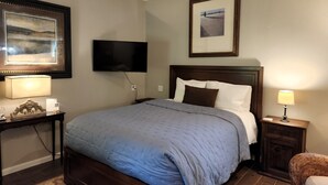 Une chambre d’hôtel avec un grand lit, un téléviseur à écran plat fixé au mur, un tableau encadré au-dessus du lit et une petite table avec une lampe.