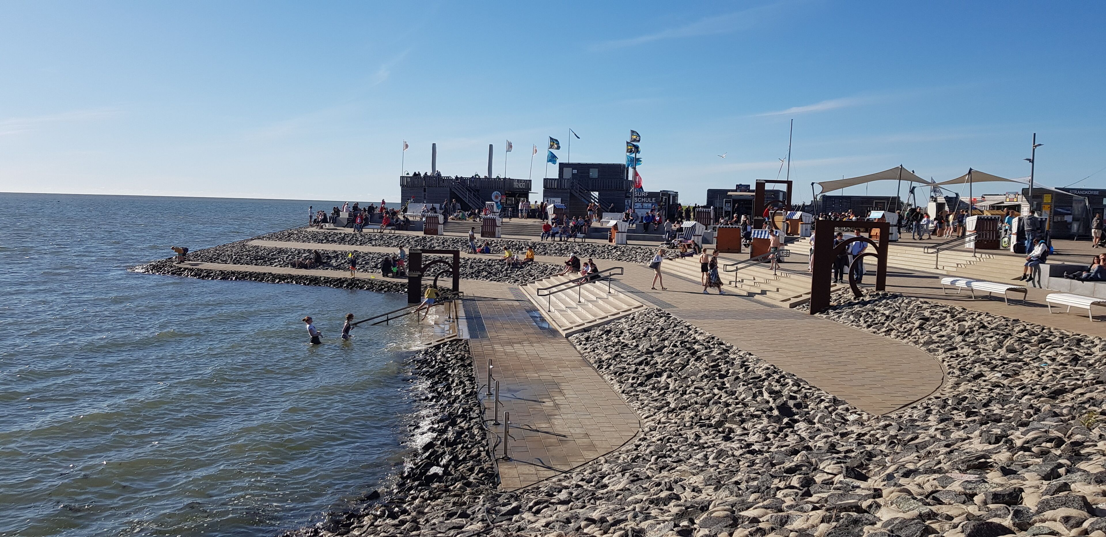 Une jetée bordée d’un rivage rocheux, une scène ornée de drapeaux, et des gens qui se promènent et qui sont assis.