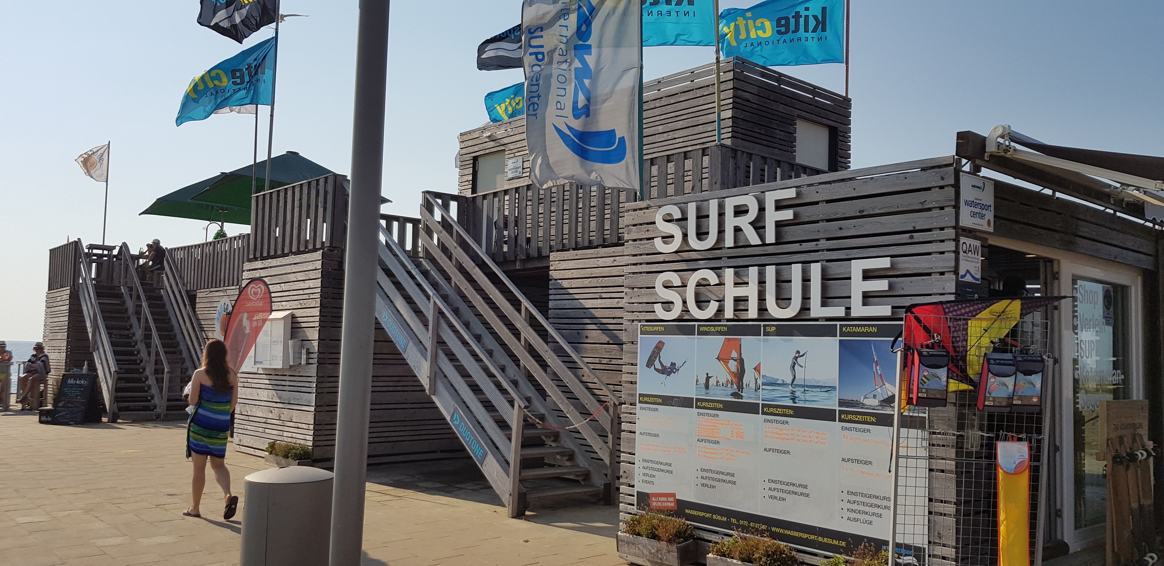 A wooden structure labeled 'SURF SCHULE' with a signboard detailing water sports courses.