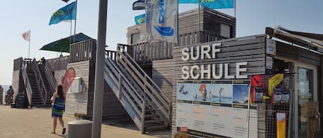 A wooden structure labeled 'SURF SCHULE' with a signboard detailing water sports courses.