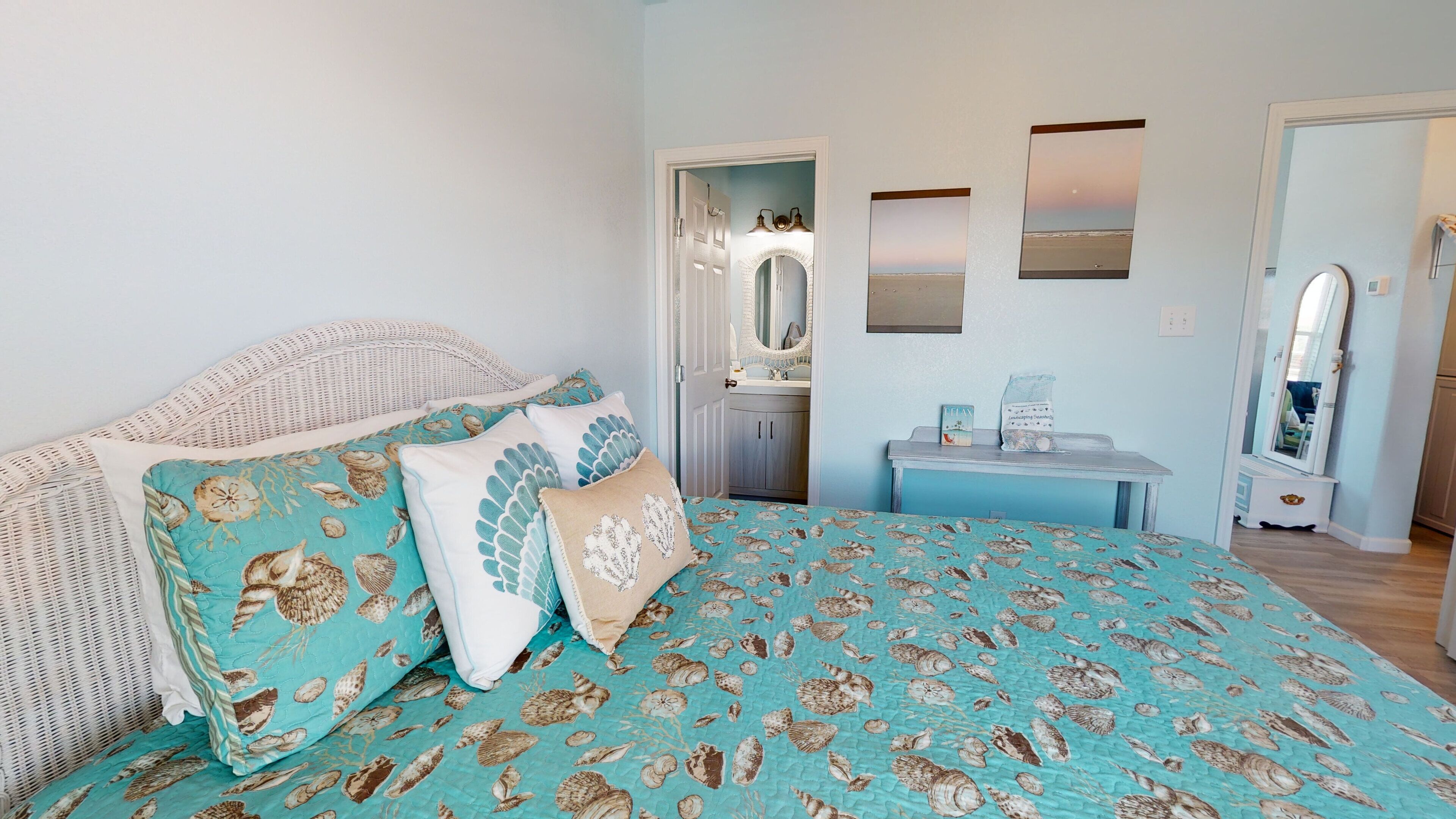A bedroom with a bed featuring a blue and white seashell patterned bedspread, a wicker headboard, and a small bedside table with a lamp and a bag of toiletries.
