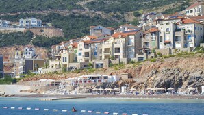 A coastal residential area with multi-story buildings, a beach with umbrellas, and a rocky hillside in the background. - Lustica Bay Marina (Radovici)