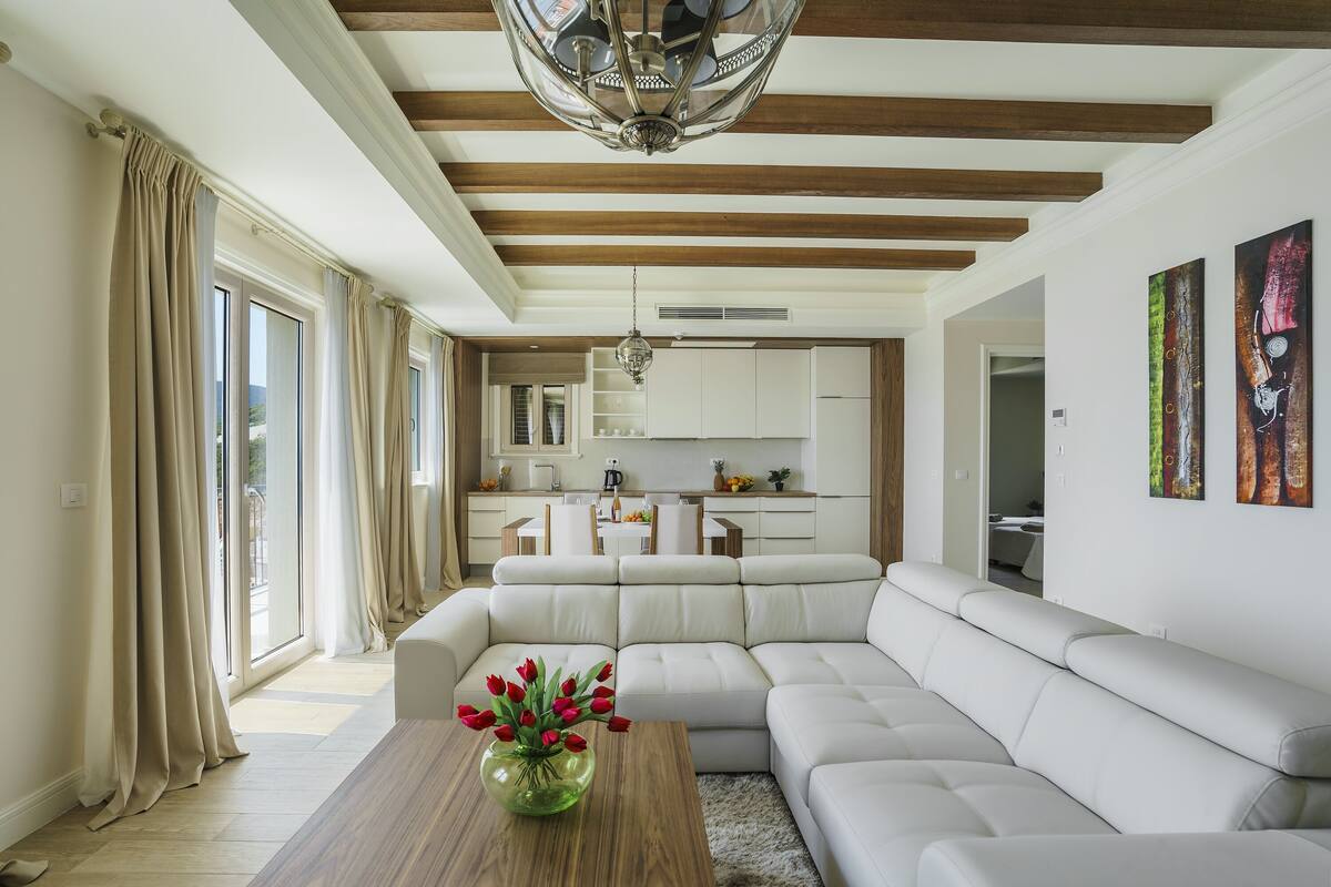 A modern living room with a sectional sofa, a wooden coffee table with a vase of red flowers, and a kitchen in the background.
