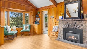 A cozy cabin interior with wooden walls, a fireplace, a bookshelf, and a window with a view of trees. - 3BR near lake w/ hot tub, views, deck, fire pit (Estes Park)