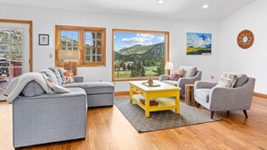 A living room with a grey sectional sofa, a grey armchair, a yellow coffee table, and a view of a mountainous landscape through large windows. - 3BR near lake w/ hot tub, views, deck, fire pit (Estes Park)