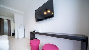 Une chambre d’hôtel moderne équipée d’un téléviseur à écran plat fixé au mur, d’un bureau avec une chaise et de deux poufs roses.