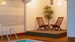 Ein Hallenbad mit klarem, blauem Wasser, zwei Holzliegen, einer Topfpflanze und einer gefliesten Wand.