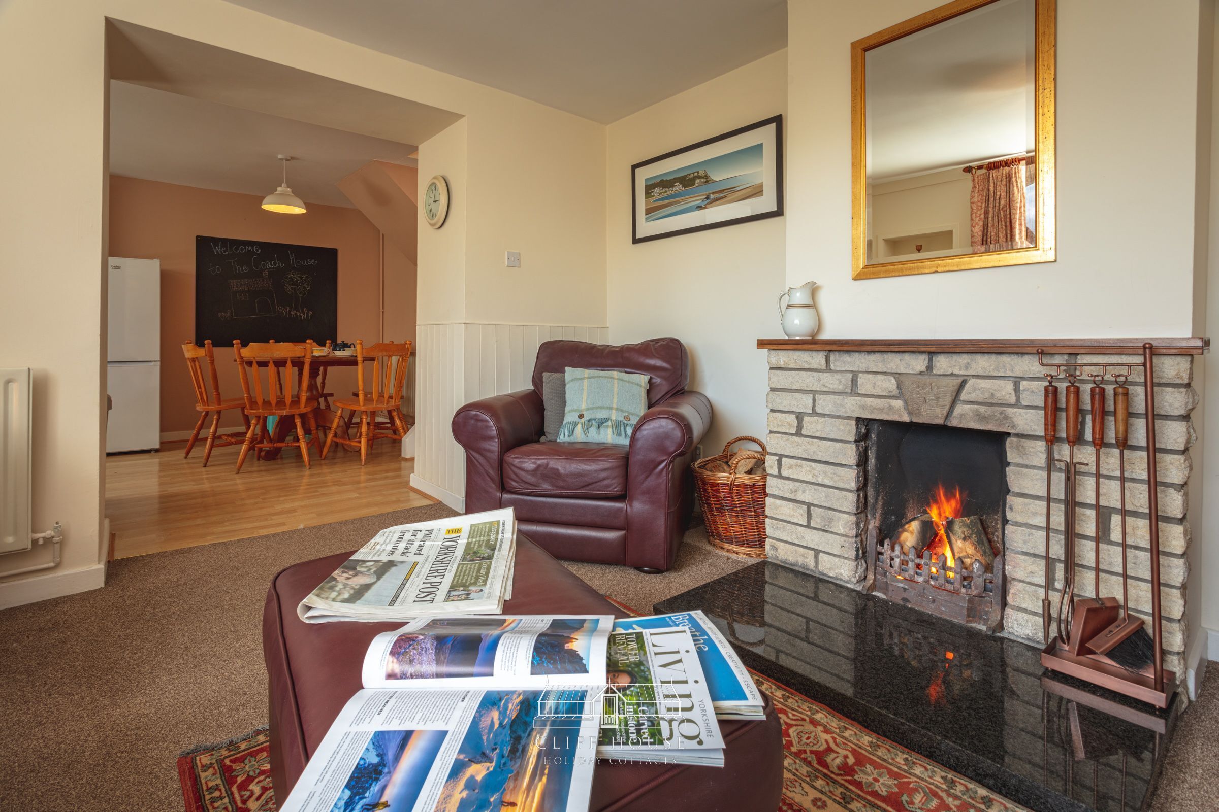 A cozy living room with a fireplace, a leather armchair, a dining area with wooden chairs, and a table with newspapers.