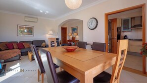 A modern living room with a sofa, dining table, and chairs. There is a kitchen visible through an open door.