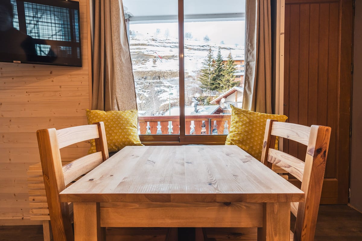 Una mesa de madera con dos sillas, una ventana que da a un paisaje nevado y un televisor montado en la pared.
