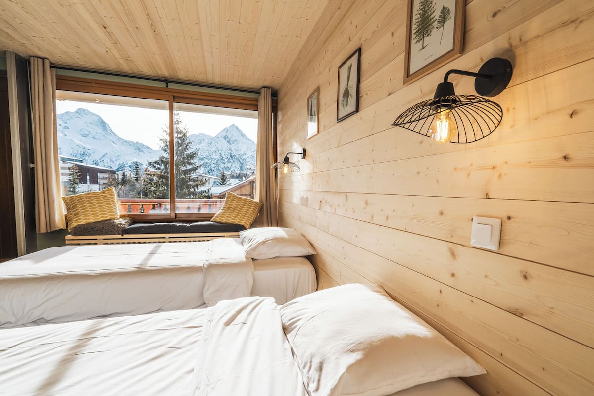 Una habitación con dos camas, paredes de madera, una ventana con vista a la montaña y una lámpara fijada a la pared.