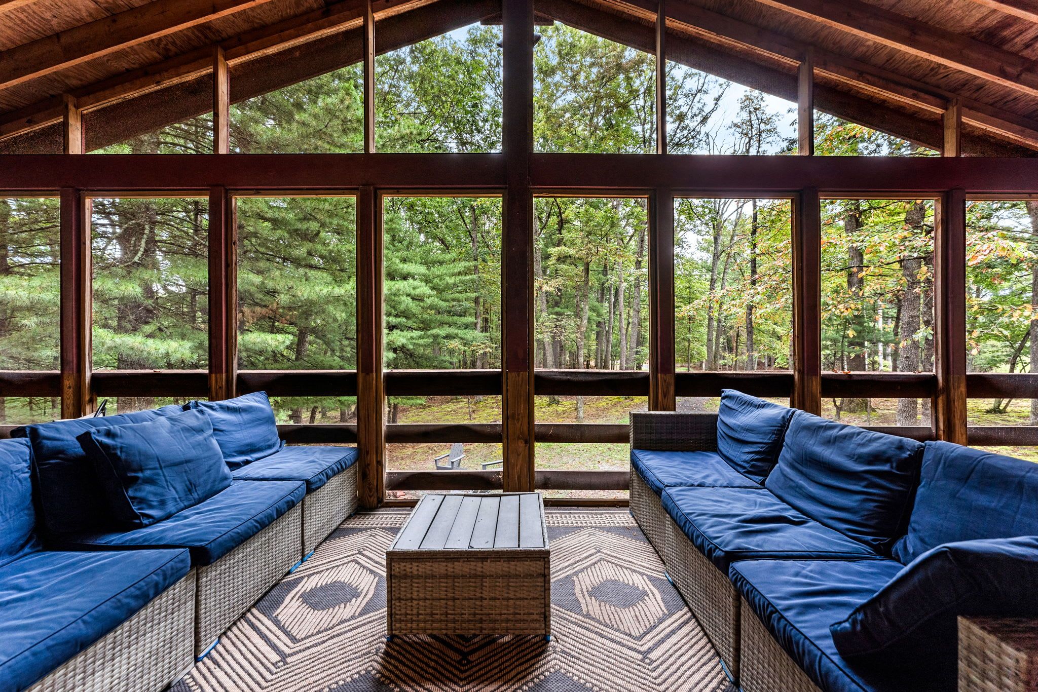 A covered outdoor seating area with wicker furniture and cushions, large windows, and a view of a forest.