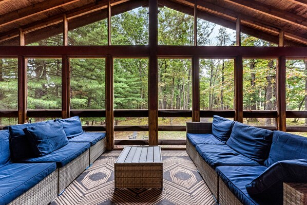 Un espacio de estar al aire libre cubierto con mobiliario de mimbre y cojines, ventanas amplias y vistas a un bosque.