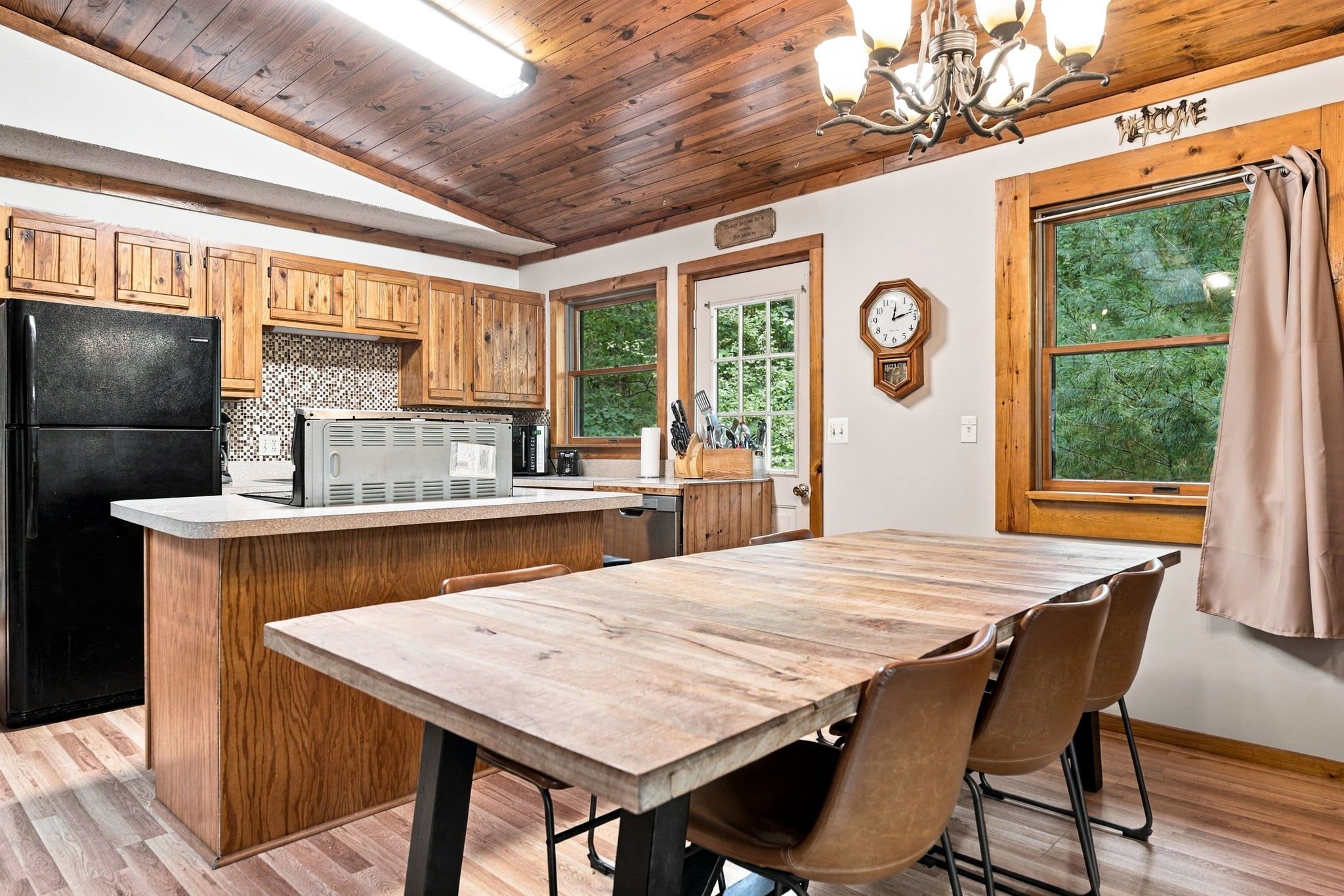 Une cuisine avec des meubles de rangement en bois, un réfrigérateur noir, une table à manger en bois avec des chaises et une horloge au mur.