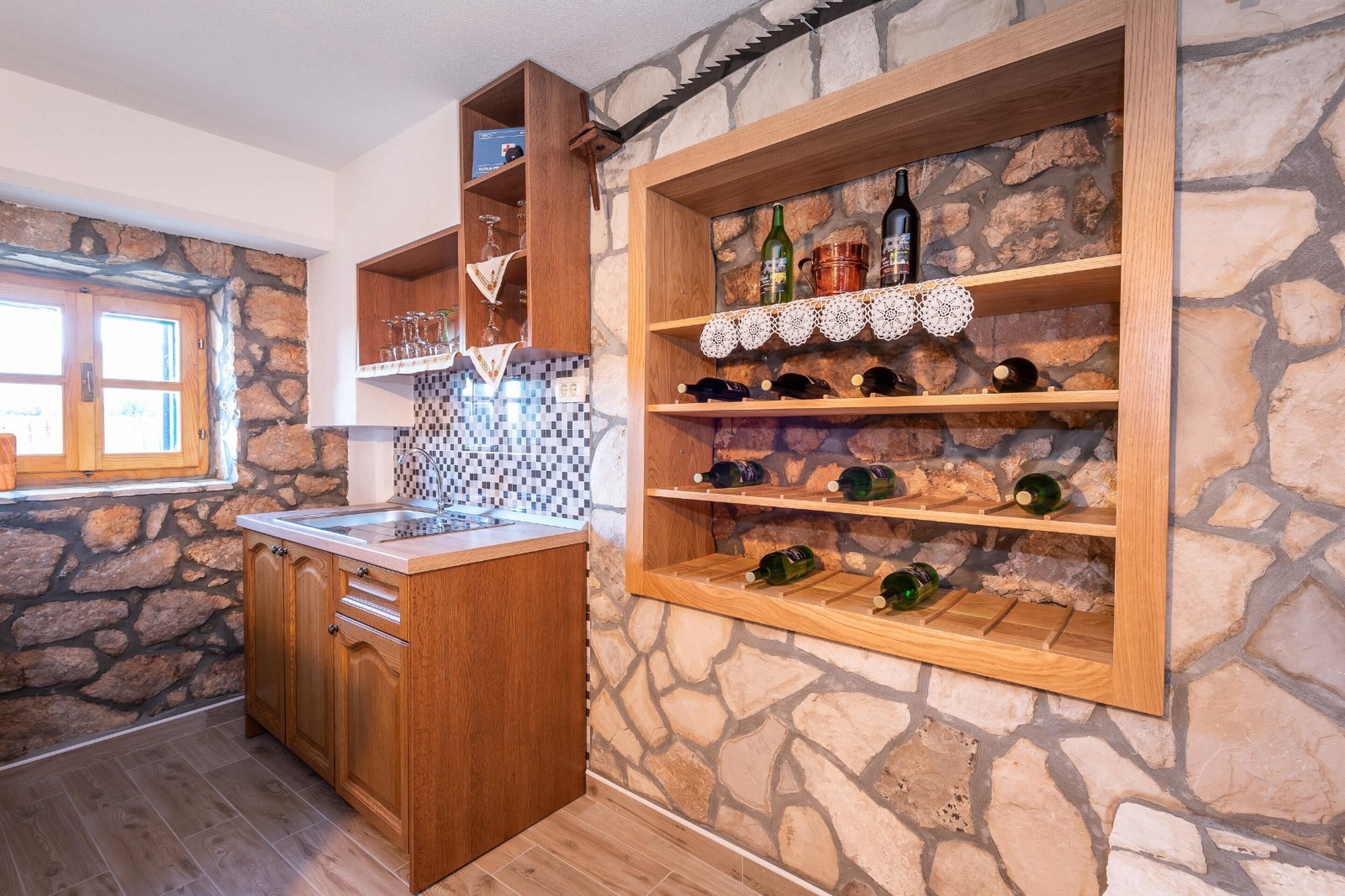 Une cuisine avec des meubles de rangement en bois, un mur en pierre et des étagères où sont disposées des bouteilles de vin et des verres.