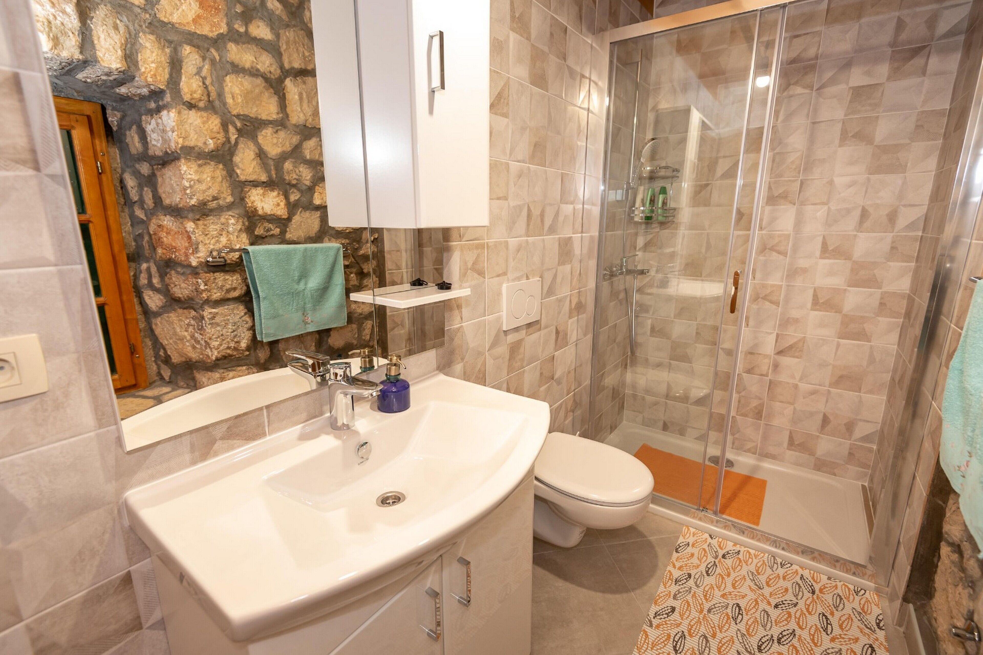 A bathroom with a stone wall, a white sink, a toilet, and a glass-enclosed shower.