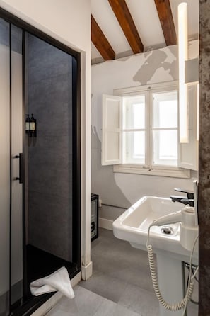 Casa de banho com cabine de duche em vidro, lavatório branco e janela com estores brancos.