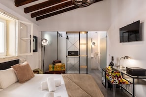 Quarto de hotel moderno com cama, mesa de cabeceira, luminária e escrivaninha com cadeira. Possui guarda-roupa com espelho e prateleira com moldura.