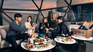A group of people sitting on a couch and at a table, enjoying a meal and drinks in a modern, glass-walled room with a view of the sky. - Mondrian Seoul Itaewon (Seoul)