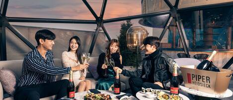 A group of people sitting on a couch and at a table, enjoying a meal and drinks in a modern, glass-walled room with a view of the sky.