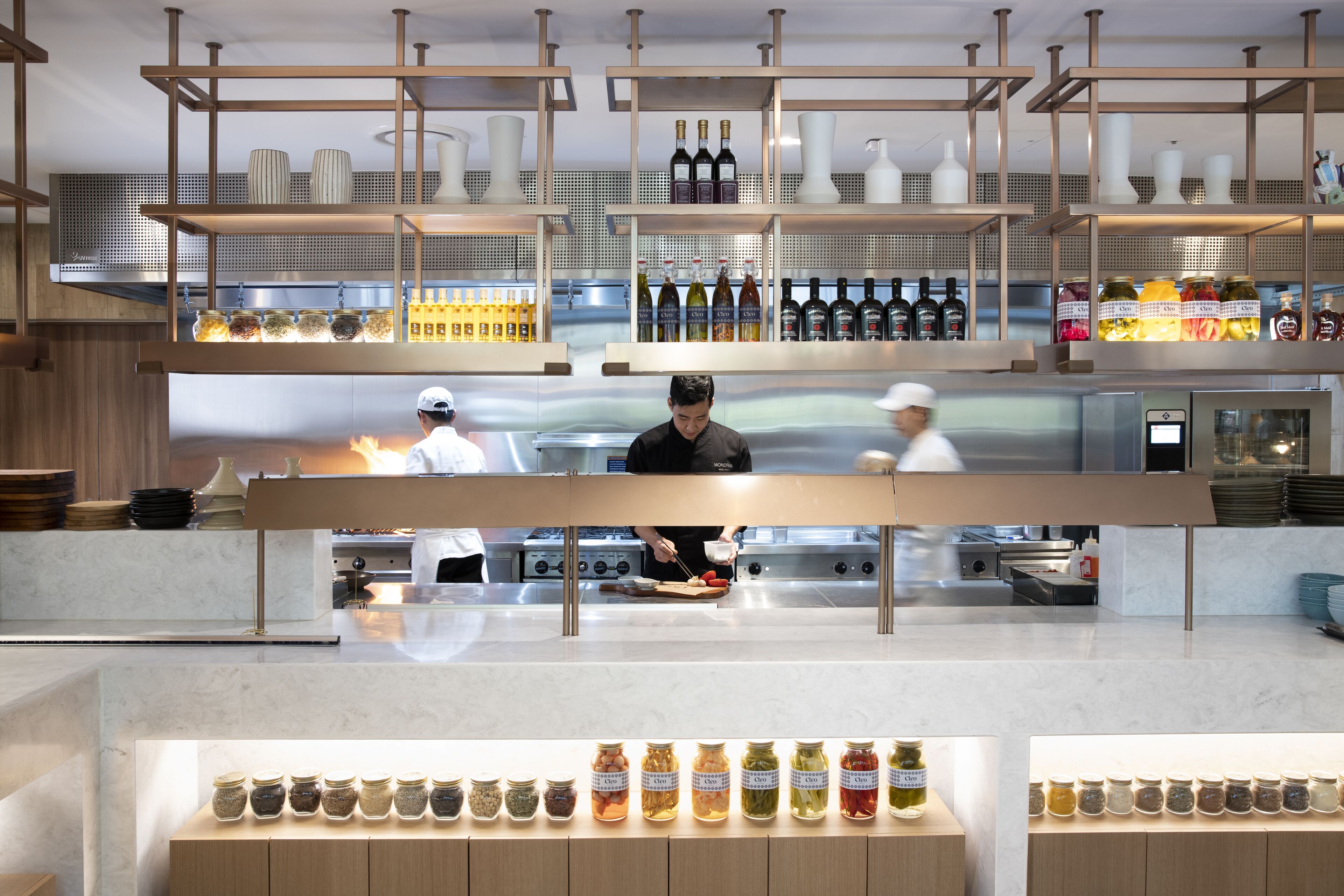 Une cuisine moderne dotée d’appareils électroménagers en acier inoxydable, d’étagères en bois où sont disposées des bouteilles et des bocaux, et d’un chef qui prépare les plats.