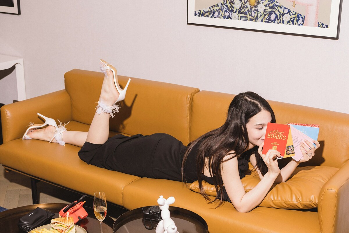 A person lying on a yellow sofa, reading a book, with a plate of food and a glass of wine on a table in front of them.