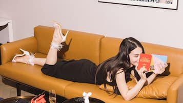 A person lying on a yellow sofa, reading a book, with a plate of food and a glass of wine on a table in front of them.