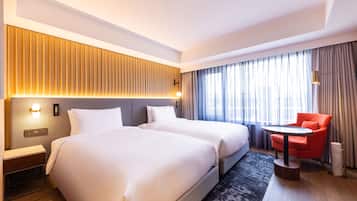Habitación de hotel con dos camas, una mesita redonda pequeña, una silla roja, una lámpara y un ventanal grande con cortinas.