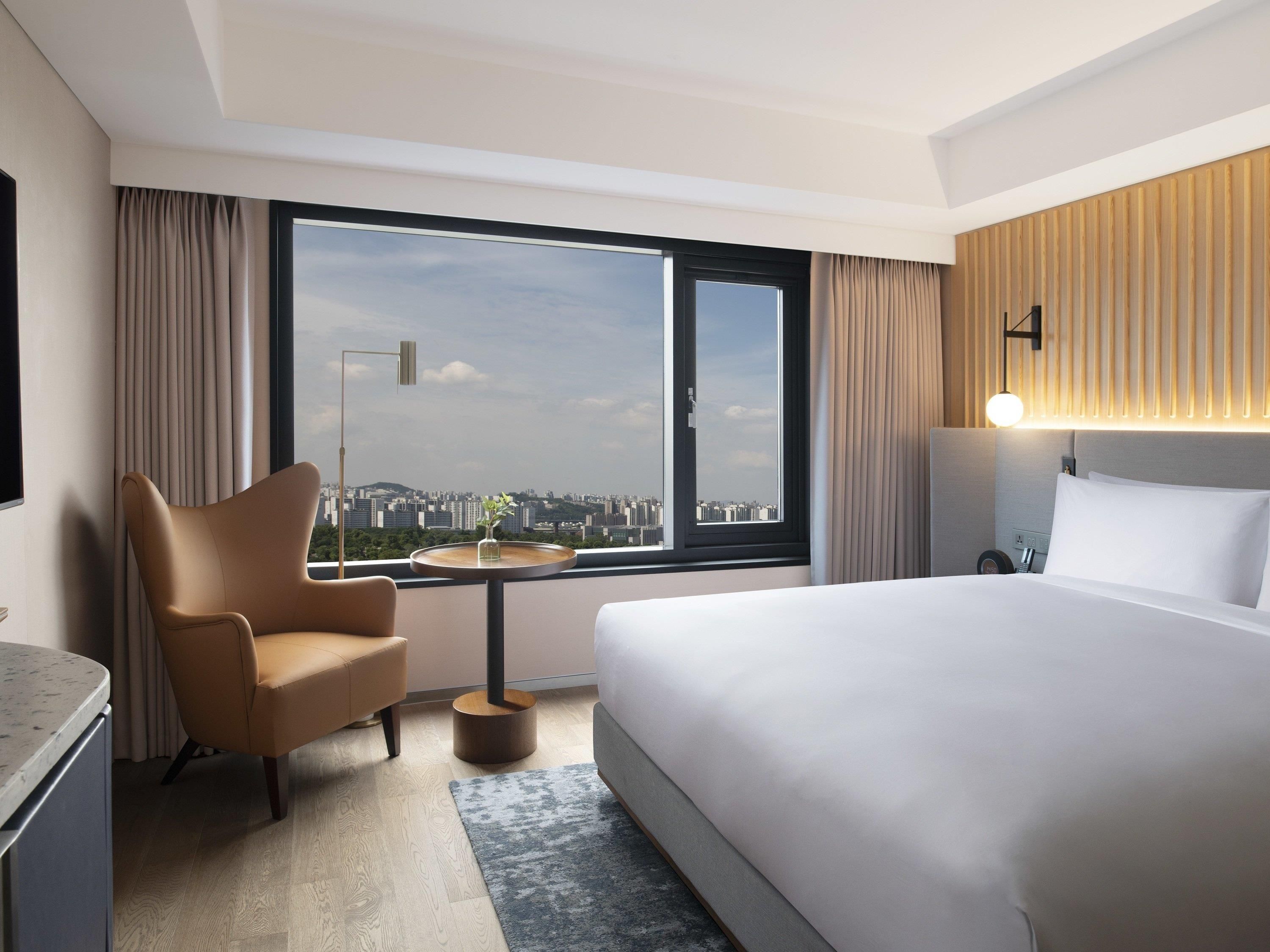 A hotel room with a bed, a chair, a small table, a lamp, and a view of the city.